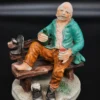Ręcznie malowana figurka – starszy mężczyzna na ławce z butelką – ceramika, wys. 20 cm